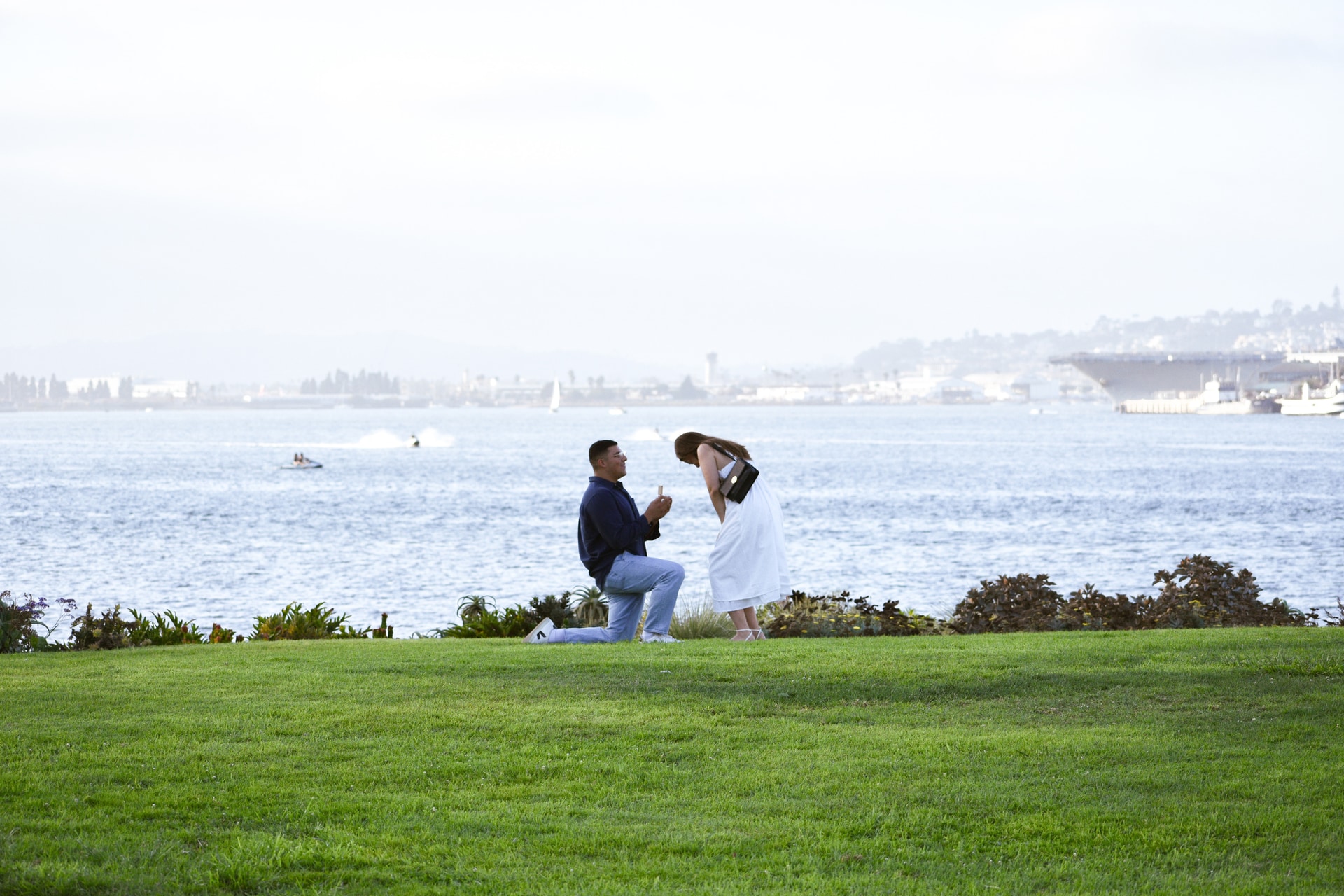 Omar on one knee holding the ring, Nati facing him, the bay behind them